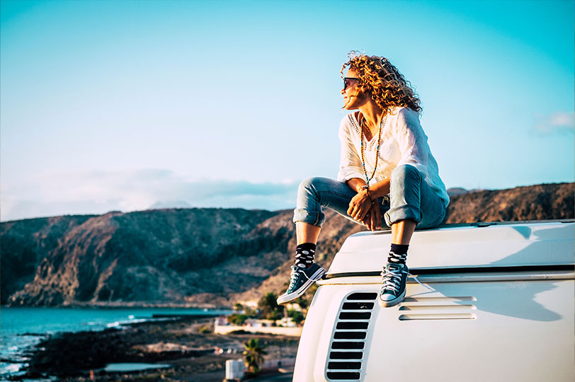 Women sat on the roof of a campervan looking out over the ocean with the sun on her face as it sets
