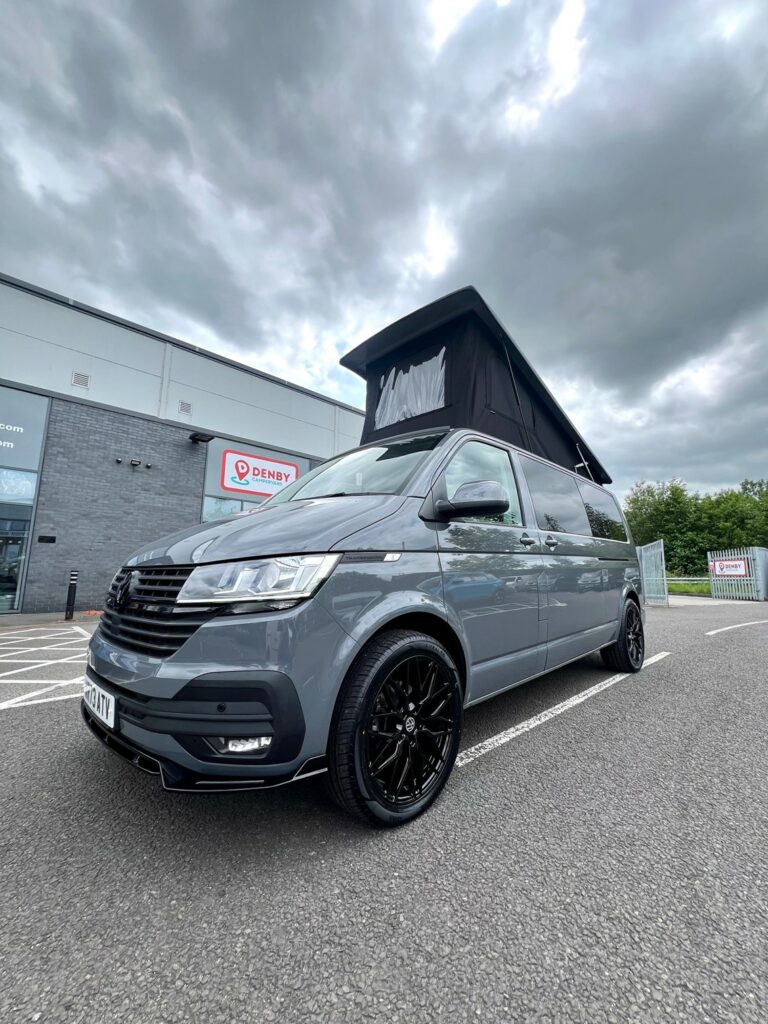 A Pure Grey Volkswagen T6.1 Transporter Highline LWB Campervan (73 Plate, DSG) with a raised pop-up roof and black alloy wheels is parked outside a modern building on a cloudy day.