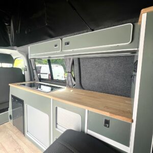 The LWB T6.1 Volkswagen Transporter Highline Campervan (DSG, Pure Grey, 73 Plate – A1439) features light green cabinets, a wooden countertop, stove, upper storage, window, textured gray walls, and minimalist light wood flooring.