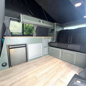 The LWB T6.1 Volkswagen Transporter Highline Campervan (DSG, Pure Grey, 73 Plate – A1439) features wood-look flooring, a compact fridge, storage cabinets, black cushioned bench seats, and small curtained windows in light gray and black tones.