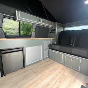 The LWB T6.1 Volkswagen Transporter Highline Campervan (DSG, Pure Grey, 73 Plate A1439) features light wood flooring, green cabinets, a compact fridge, window with gray curtains, and a black cushioned bench in a clean, organized interior.