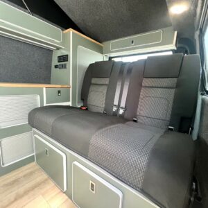 The LWB T6.1 Volkswagen Transporter Highline Campervan (DSG, Pure Grey, 73 Plate, A1439) features a grey and black upholstered bench seat with seat belts, under-seat storage, sleek cabinetry, and a light wood floor.