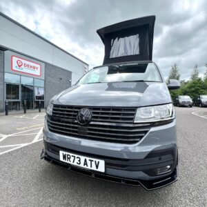 A Pure Grey Volkswagen Transporter T6.1 Highline LWB campervan (DSG, 73 Plate, A1439) with its pop-up roof extended is parked outside a Denby building on a cloudy day, with several other vehicles visible in the background.