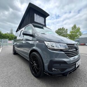 A Pure Grey LWB Volkswagen Transporter T6.1 Highline Campervan (DSG, 73 Plate, A1439) with a raised black pop-top roof is parked in a lot on an overcast day, with trees and industrial buildings in the background.