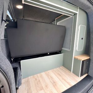 The LWB T6.1 Volkswagen Transporter Highline Campervan (DSG, Pure Grey, 73 Plate – A1439) features a clean, minimal interior with light wood flooring, a black sofa bed, grey-green walls, wood-topped counter, and storage cabinets.