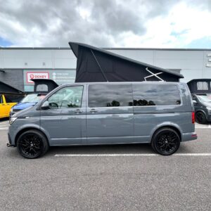 A Pure Grey LWB Volkswagen Transporter Highline Campervan (T6.1, DSG, 73 Plate, A1439) with a pop-up roof, black wheels, and tinted windows is parked in a lot outside a building under a cloudy sky.