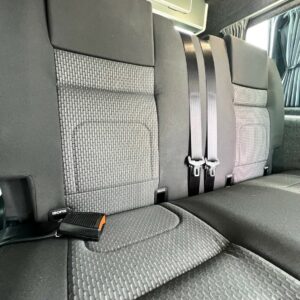 Close-up of the back seat in the LWB T6.1 Volkswagen Transporter Highline Campervan (DSG, Pure Grey, 73 Plate, A1439), showing dark gray patterned fabric upholstery with visible seatbelt buckles and two fastened seatbelts in the center section.