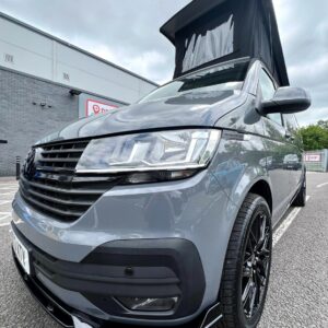 A Pure Grey LWB Volkswagen Transporter T6.1 Highline Campervan (73 Plate, DSG, A1439) with an elevated pop-up roof is parked on an asphalt lot near a modern building; the low-angle shot highlights its front and raised roof.