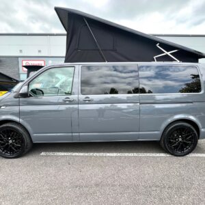 A LWB T6.1 Volkswagen Transporter Highline Campervan DSG in Pure Grey (73 Plate, A1439) with a raised black pop-up roof, black wheels, and tinted windows is parked in a lot with other vehicles and a building visible in the background.
