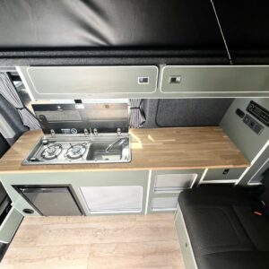 The LWB T6.1 Volkswagen Transporter Highline Campervan (DSG, Pure Grey, 73 Plate – A1439) features a modern kitchen with wood countertop, two-burner stove, mini fridge, sink, storage cabinets, and a black cushioned seat.