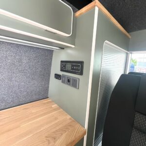 The LWB - T6.1 Volkswagen Transporter Highline Campervan (DSG, Pure Grey, 73 Plate, A1439) features a compact interior with a wooden countertop, green cabinetry, control panel, power outlets, and grey fabric wall beside a seat and sunlit window.