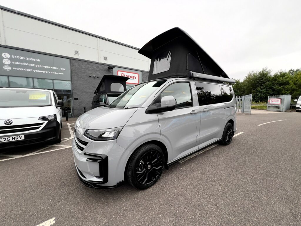 A 2025 ALL-NEW Volkswagen Transporter T7 DSG Auto Commerce Plus Campervan in Stone Grey (Deluxe SWB, Stock A1393) with a raised pop-top roof, black alloy wheels, and tinted windows is parked outside a building.