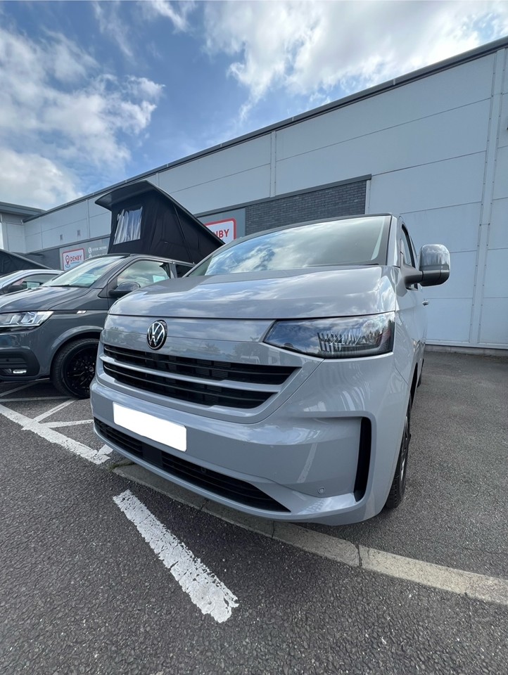 A Stone Grey Deluxe SWB - 2025 ALL-NEW Volkswagen Transporter T7 DSG Auto Commerce Plus Campervan (Stock A1393) is parked in a lot beside another vehicle, with a building and partly cloudy sky in the background. Front and side are visible; plate is blurred.