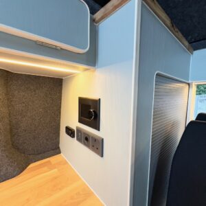 Deluxe SWB - 2025 Volkswagen Transporter T7 DSG Auto Commerce Plus Campervan in Stone Grey (Stock A1393) features a modern interior with light wood countertop, white cabinetry, wood trim, built-in outlets, textured grey wall panel, and under-cabinet lighting.