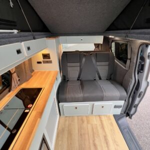 The Deluxe SWB 2025 Volkswagen Transporter T7 Campervan in Stone Grey features a wooden floor, left-side kitchen with stove and cabinets, gray bench seat with seatbelts at the rear, and dark fabric-lined ceiling. Stock A1393.