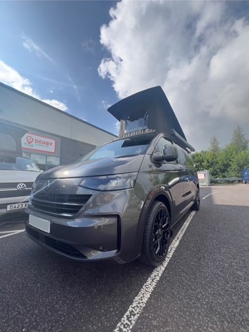 A graphite Deluxe SWB 2025 Volkswagen Transporter DSG Auto Commerce Plus Campervan (Stock A1391) with pop-up roof is parked next to another vehicle outside a Denby building, trees and a partly cloudy sky in the background.