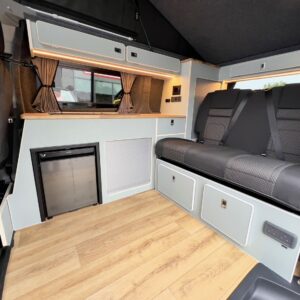 The Deluxe SWB - 2025 ALL-NEW Volkswagen Transporter T7 DSG Auto Commerce Plus Campervan (Stone Grey, Stock A1393) features wood flooring, a small fridge, light gray cabinets, overhead storage, cushioned seatbelts seats, and brown curtains.