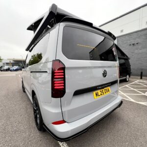 A Stone Grey 2025 ALL-NEW Volkswagen Transporter T7 DSG Auto Commerce Plus Campervan (Deluxe SWB, Stock A1393) with black rims is parked near a disabled bay, rear hatch open, UK plate ML25 OVA. Building and cars seen behind.