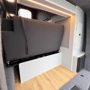 Interior of the Deluxe SWB - 2025 ALL-NEW Volkswagen Transporter T7 DSG Auto Commerce Plus Campervan in Stone Grey (Stock A1393) featuring a raised black sofa bed, white storage, wood-look flooring, and modern grey-white paneling with soft lighting.