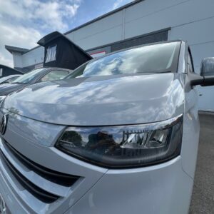 Close-up of the front of a 2025 ALL-NEW Volkswagen Transporter T7 DSG Auto Commerce Plus Campervan in Stone Grey (Stock A1393), reflecting sky and clouds, with other vehicles and a modern industrial building in the background.