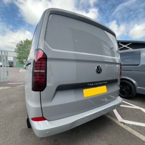A Stone Grey 2025 ALL-NEW Volkswagen Transporter T7 DSG Auto Commerce Plus Campervan (Deluxe SWB, Stock A1393) is parked in a lot with its rear facing the camera. The sky is partly cloudy and another van is beside it. License plate is blurred for privacy.