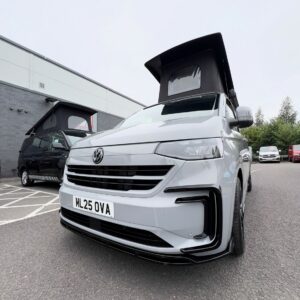 A Stone Grey Deluxe SWB 2025 ALL-NEW Volkswagen Transporter T7 DSG Auto Commerce Plus Campervan (Stock A1393) is parked on a paved lot. Other vehicles, including another camper van, appear in the background. License plate: ML25 OVA.