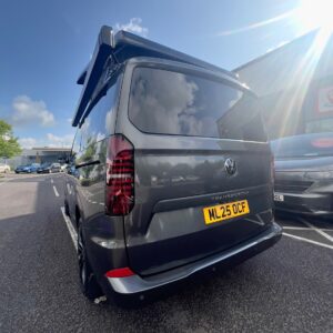 A 2025 ALL-NEW Volkswagen Transporter DSG Auto Commerce Plus Campervan in Graphite Dust (Stock A1391) with a raised pop-up roof is parked in a lot, license plate ML25 OCF, with other cars and a building nearby under a partly cloudy sky.