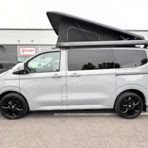 A stone grey Deluxe SWB 2025 Volkswagen Transporter T7 DSG Auto Commerce Plus Campervan (Stock A1393) with a black pop-up roof, black wheels, and tinted windows is parked outside a building, with other vehicles and a Denby sign in view.