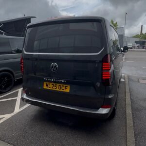 A 2025 ALL-NEW Volkswagen Transporter DSG Auto Commerce Plus Campervan in Graphite Dust (Deluxe SWB, Stock A1391) is parked in a lot next to another vehicle, with buildings visible in the background on a cloudy day.