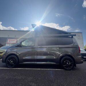A Deluxe SWB 2025 ALL-NEW Volkswagen Transporter DSG Auto Commerce Plus Campervan in Graphite Dust (Stock A1391) with its pop-up roof open is parked under a sunny, cloud-dotted sky, sunlight creating a dramatic effect behind it.