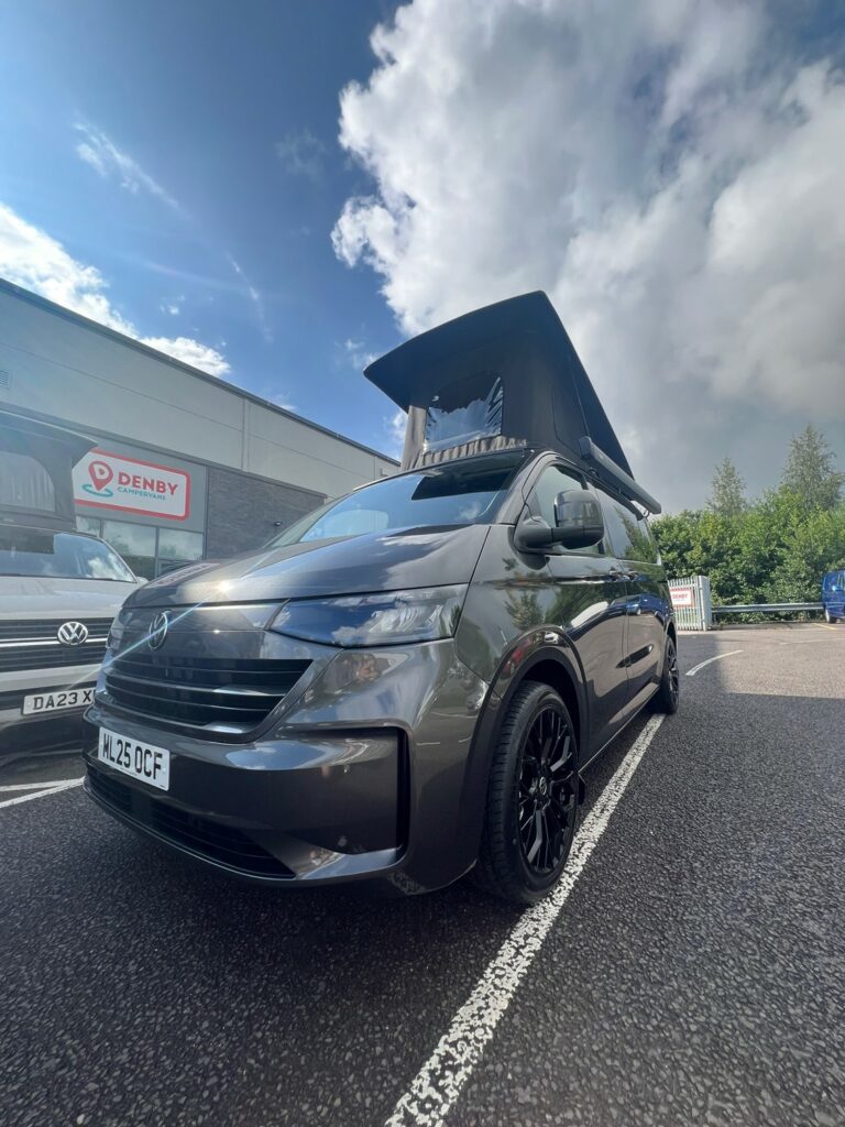A 2025 ALL-NEW Volkswagen Transporter DSG Auto Commerce Plus Campervan in Graphite Dust (Stock A1391) with its pop-up roof extended is parked outside a building under a partly cloudy sky, featuring black wheels and surrounded by other vans.