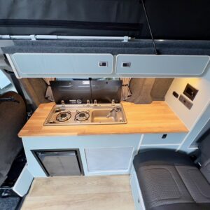 The Deluxe SWB - 2025 Volkswagen Transporter T7 DSG Auto Commerce Plus Campervan in Stone Grey (Stock A1393) features a wooden countertop, two-burner stove, sink, gray cabinets, dual seats, and light wood flooring.
