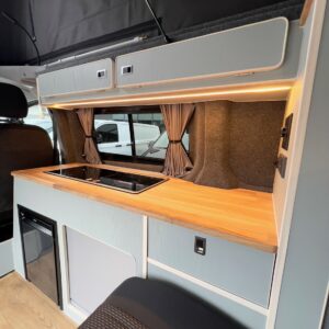 The Deluxe SWB - 2025 ALL-NEW Volkswagen Transporter T7 DSG Auto Commerce Plus Campervan in Stone Grey (Stock A1393) features a compact, modern interior with light wood countertop, built-in stove, pale blue cabinets, small fridge, and taupe curtains.
