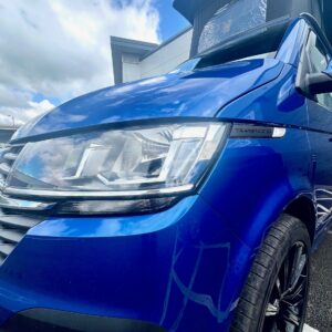 A 2023 T6.1 VW Transporter (110 BHP, manual) in Ravenna Blue (A1459) with a raised pop-top roof is parked outside on a cloudy day, its shiny surface reflecting the sky and surroundings.