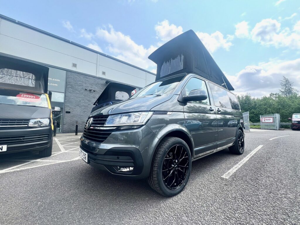 An Indium Grey SWB T6.1 Volkswagen Transporter Highline Campervan (73 plate, A1438) is parked outside a dealership on a sunny day, with another similar campervan displayed for sale beside it.