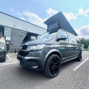 An Indium Grey SWB T6.1 Volkswagen Transporter Highline Campervan (73 plate, A1438) is parked outside a dealership on a sunny day, with another similar campervan displayed for sale beside it.