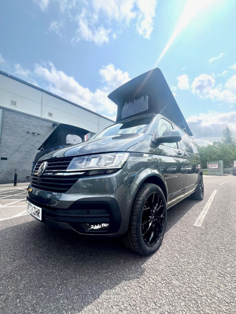 An Indium Grey SWB T6.1 Volkswagen Transporter Highline Campervan (73 plate, A1438) is parked in a lot beside a modern building under partly cloudy skies. The low-angle photo highlights the vehicles front and side with pop-top roof raised.