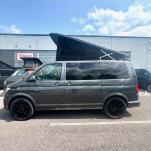 An Indium Grey SWB T6.1 Volkswagen Transporter Highline Campervan (73 plate, A1438) with a pop-up roof is parked outside a building under a blue sky, with part of another vehicle visible behind it.