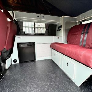 The SWB T6.1 Volkswagen Transporter Highline Campervan in Fortana Red (23 Plate, A1443) features red quilted seats, a small fridge, white storage cabinets, and a dark speckled floor—compact, clean, and ideal for travel or camping.