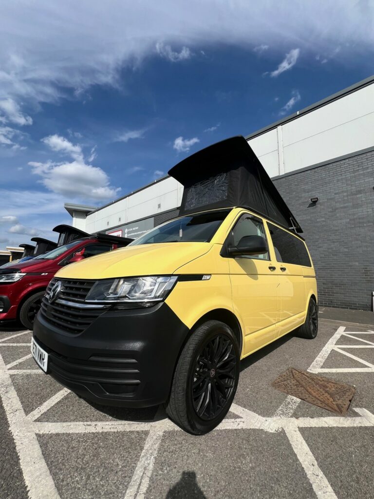 A Sunny Yellow SWB Volkswagen T6.1 Transporter Startline Campervan (71 Plate, A1441) with a pop-up roof is parked in a marked parking space outside a building under a partly cloudy sky.