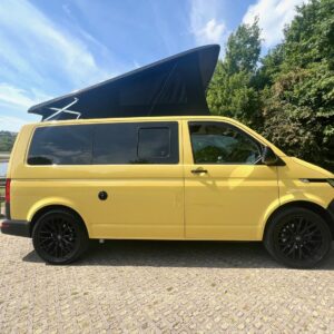 A Sunny Yellow SWB Volkswagen T6.1 Transporter Startline Campervan (71 Plate, A1441) with a black pop-up roof is parked on cobblestones near trees, under a partly cloudy blue sky.