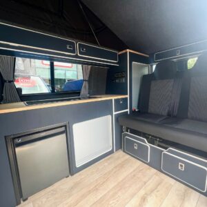 The SWB T6.1 Volkswagen Transporter Highline Campervan (Indium Grey, 73 plate, A1438) features a compact kitchenette with fridge, cabinets, and sink next to a cushioned bench seat. Light wood flooring contrasts with dark cabinets and walls; curtains cover the windows.