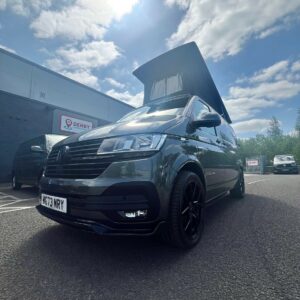 An Indium Grey SWB T6.1 Volkswagen Transporter Highline Campervan (73 plate, A1433) is parked on asphalt under a partly cloudy sky, with a building and several cars in the background. The front plate reads WG73 MRY.