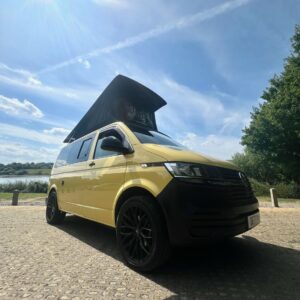 A SWB T6.1 Volkswagen Transporter Startline Campervan in Sunny Yellow (71 Plate, A1441) with a pop-top roof is parked on cobblestone near a lake, surrounded by trees under a blue sky with wispy clouds.