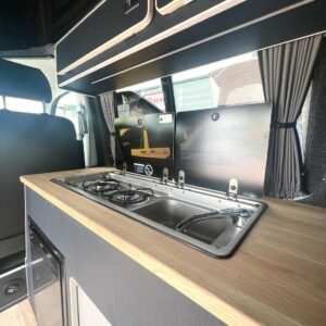 The image shows the SWB T6.1 Volkswagen Transporter Highline Campervan (Indium Grey, 73 plate, A1438) with a wooden countertop kitchen featuring a two-burner stovetop and sink, overhead cabinets, small fridge below, and curtained window above.