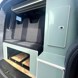 The SWB T6.1 Volkswagen Transporter Highline Campervan (Indium Grey, 73 plate, A1433) features a modern interior with two black/gray seats, teal storage cabinets, and a wooden pull-out platform for compact travel or camping functionality.