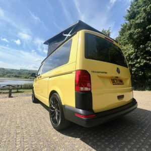 A SWB T6.1 Volkswagen Transporter Startline Campervan in Sunny Yellow (71 Plate, A1441) is parked on paving near a lake and trees on a sunny day.