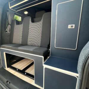 The SWB T6.1 Volkswagen Transporter Highline Campervan (Indium Grey, 73 plate, A1438) features a modern interior with three black cushioned seats, light wood flooring, blue cabinetry with white trim, overhead shelf, and curtain above the seats.