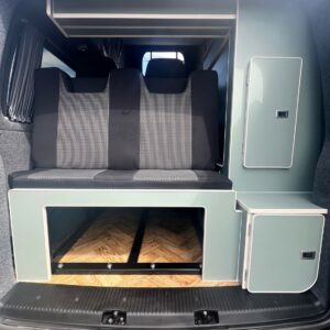 The image features the rear interior of the SWB - T6.1 Volkswagen Transporter Highline Campervan (Indium Grey, 73 plate, A1433) with a black bench seat, green cabinets, wood-patterned floor, and organized under-seat storage.