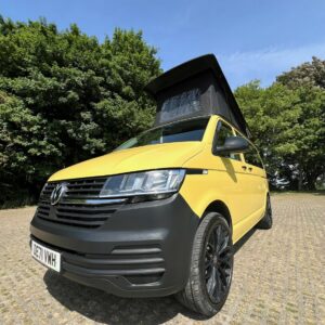 A SWB T6.1 Volkswagen Transporter Startline Campervan in Sunny Yellow (71 Plate, A1441) is parked on a paved surface near green trees under a clear blue sky.